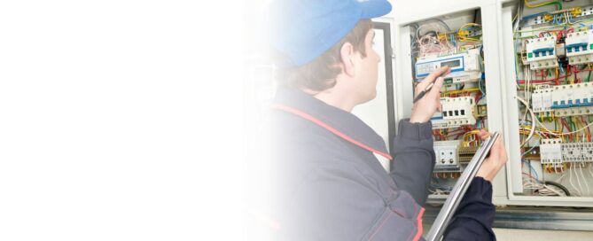 Técnico electricista sonriente con overol azul y casco, usando un multímetro para medir el voltaje en un panel de control eléctrico industrial.