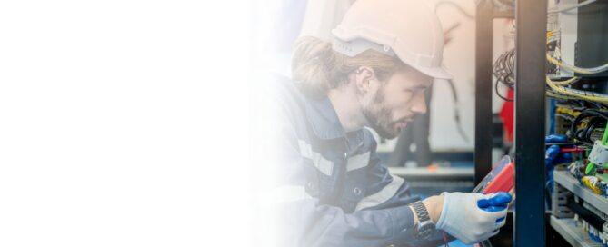 Electricista industrial sonriente con overol azul y casco amarillo, usando un multímetro para medir voltaje en un panel de control eléctrico industrial.